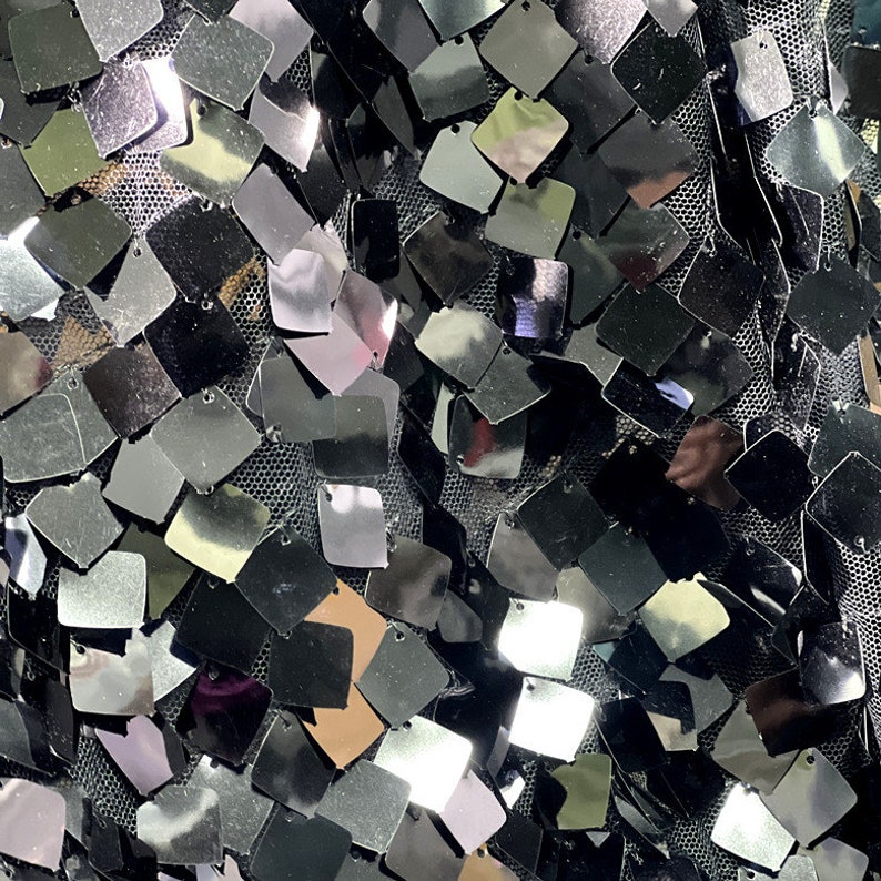 May include: Close-up of a fabric covered in small, square, silver sequins. The sequins are attached to a black mesh backing. The sequins reflect light, creating a shimmering effect. The overall texture is dense and visually striking.