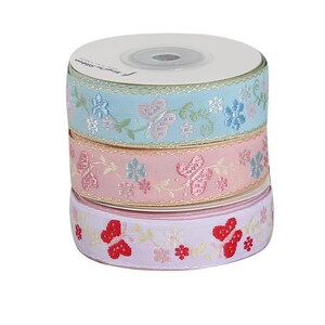 May include: A stack of three spools of decorative ribbon. The top spool is light blue with embroidered butterflies and flowers. The middle spool is pink, and the bottom spool is lavender with red butterflies.