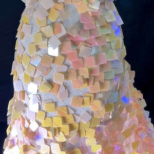 May include: Close-up of a garment covered in iridescent square sequins. The sequins are in shades of yellow, pink, and white, and are attached to a white mesh fabric. The background is dark.