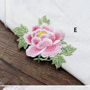 Op de afbeelding: Een geborduurde bloem in roze en wit met groene bladeren. De bloem bevindt zich op een witte stoffen achtergrond met de letter "E" in de rechterbovenhoek.