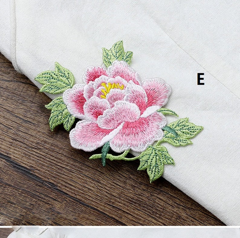 Op de afbeelding: Een geborduurde bloem in roze en wit met groene bladeren. De bloem bevindt zich op een witte stoffen achtergrond met de letter "E" in de rechterbovenhoek.
