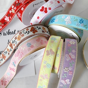 May include: A collection of decorative ribbons in various colors, including red, white, blue, pink, yellow, and purple. Each ribbon features floral and butterfly patterns. The ribbons are spooled on white cardboard rolls.