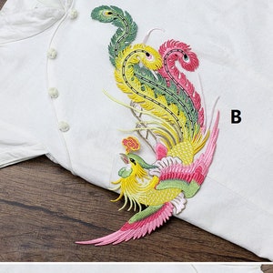 Op de afbeelding: Een kleurrijk geborduurd pleister met een feniks met roze, gele en groene veren. De pleister is op een witte stoffen achtergrond. De letter "B" is zichtbaar in de rechterbovenhoek van de afbeelding.