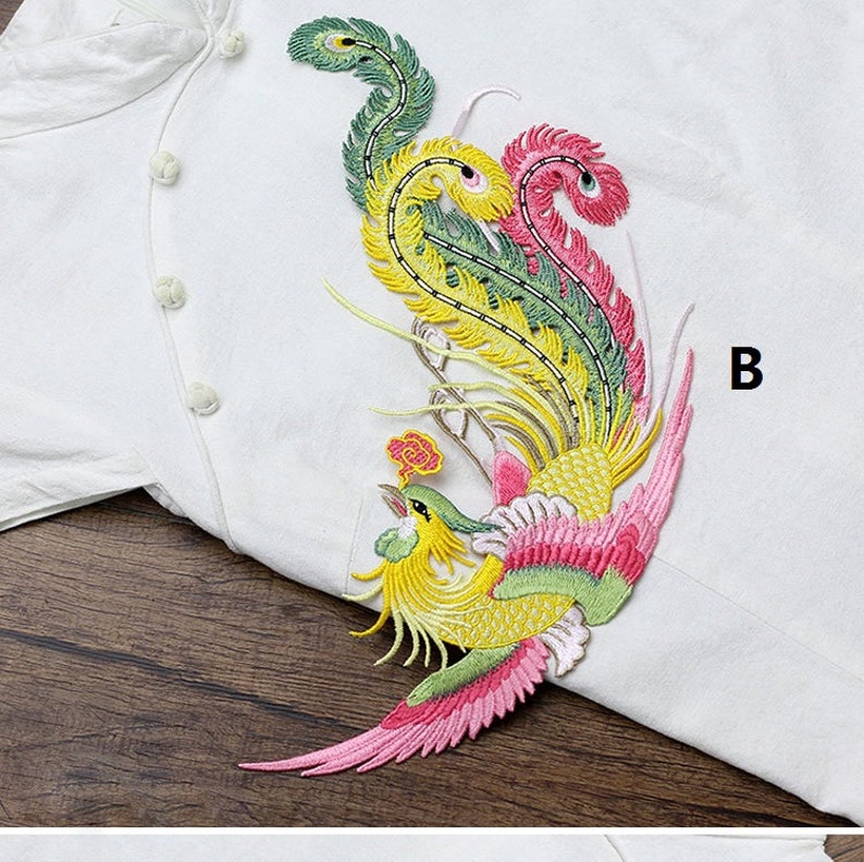 Op de afbeelding: Een kleurrijk geborduurd pleister met een feniks met roze, gele en groene veren. De pleister is op een witte stoffen achtergrond. De letter "B" is zichtbaar in de rechterbovenhoek van de afbeelding.