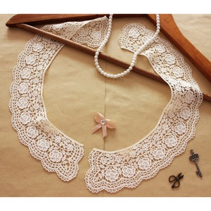 May include: Ivory lace collar with floral detailing, displayed on a wooden hanger. A pearl necklace, a pink bow with a rhinestone, a vintage key, and small scissors are also present. The collar is a delicate accessory.