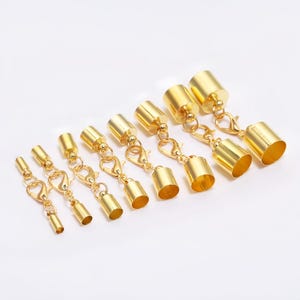May include: A collection of gold-colored jewelry clasps and end caps. The clasps have a lobster claw design, and the end caps are cylindrical. The jewelry findings are arranged on a white surface.