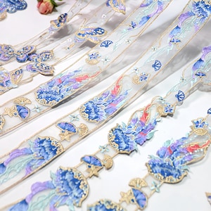 以下が含まれることがあります： 青と紫の花と蝶のデザインが繰り返される繊細な刺繍リボン。リボンは、金色のトリムが付いた透明な白い背景で、エレガントで華やかな美しさを生み出しています。クラフトや装飾に最適です。