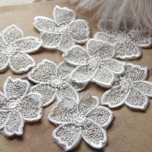 May include: A collection of white, embroidered flower appliques. Each applique features a five-petal design with intricate lace-like detailing. The appliques are scattered on a neutral-toned surface, suggesting a craft or sewing project.