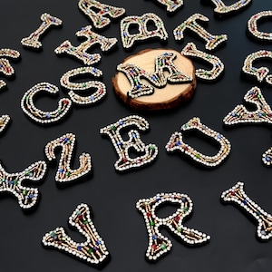 May include: Scattered across a dark surface are various beaded letters in a variety of colors. The letters are outlined in black and have a gold-colored border. Some letters are on a small wooden disc.