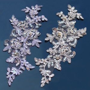 May include: Two white lace appliques featuring silver sequins and pearl beads. The appliques are shaped like floral branches with leaves and flowers.