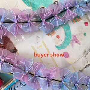 May include: A garland of sheer, pastel-colored bows, transitioning from lavender to blue, is strung along a black cord. Each bow is accented with a small, white bead. The words "buyer show" are visible in the image.
