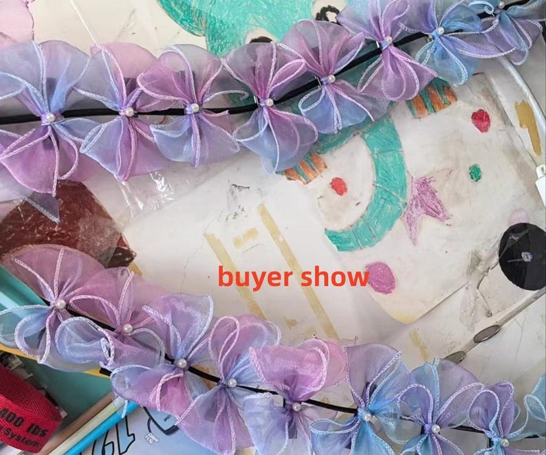 May include: A garland of sheer, pastel-colored bows, transitioning from lavender to blue, is strung along a black cord. Each bow is accented with a small, white bead. The words "buyer show" are visible in the image.