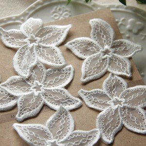 Op de afbeelding: Vijf witte kantbloem applicaties met een delicaat bloemenpatroon. De applicaties zijn gemaakt van een transparante stof en hebben een licht verhoogde textuur.