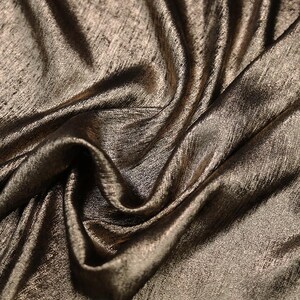 Pode incluir: Close-up de um tecido dourado met&aacute;lico com um padr&atilde;o texturizado sutil. O tecido est&aacute; drapeado e dobrado, mostrando sua textura macia e fluida.