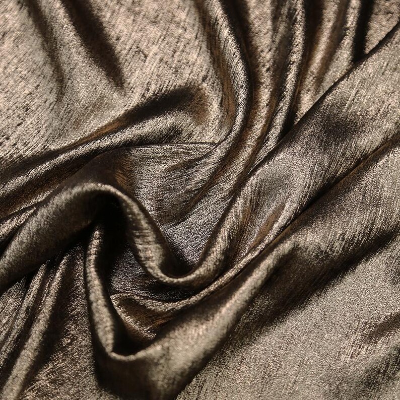Pode incluir: Close-up de um tecido dourado met&aacute;lico com um padr&atilde;o texturizado sutil. O tecido est&aacute; drapeado e dobrado, mostrando sua textura macia e fluida.