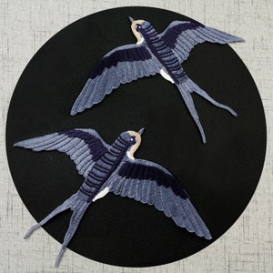 May include: Two blue and black embroidered swallow bird patches on a black background.