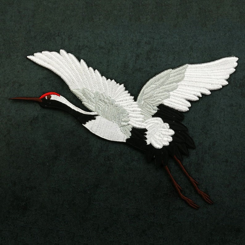 May include: White embroidered crane with black and red accents, flying with wings spread wide against a dark green background.
