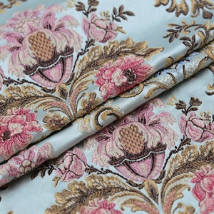 May include: Light blue fabric with a floral pattern. The design features large pink and brown flowers with detailed leaves and stems. The fabric is folded, showing the texture and sheen of the material.