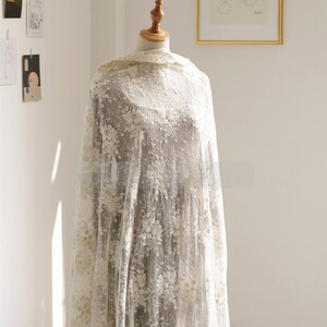 May include: A white lace fabric with a delicate floral pattern. The fabric is draped over a mannequin and has a soft, flowing texture.