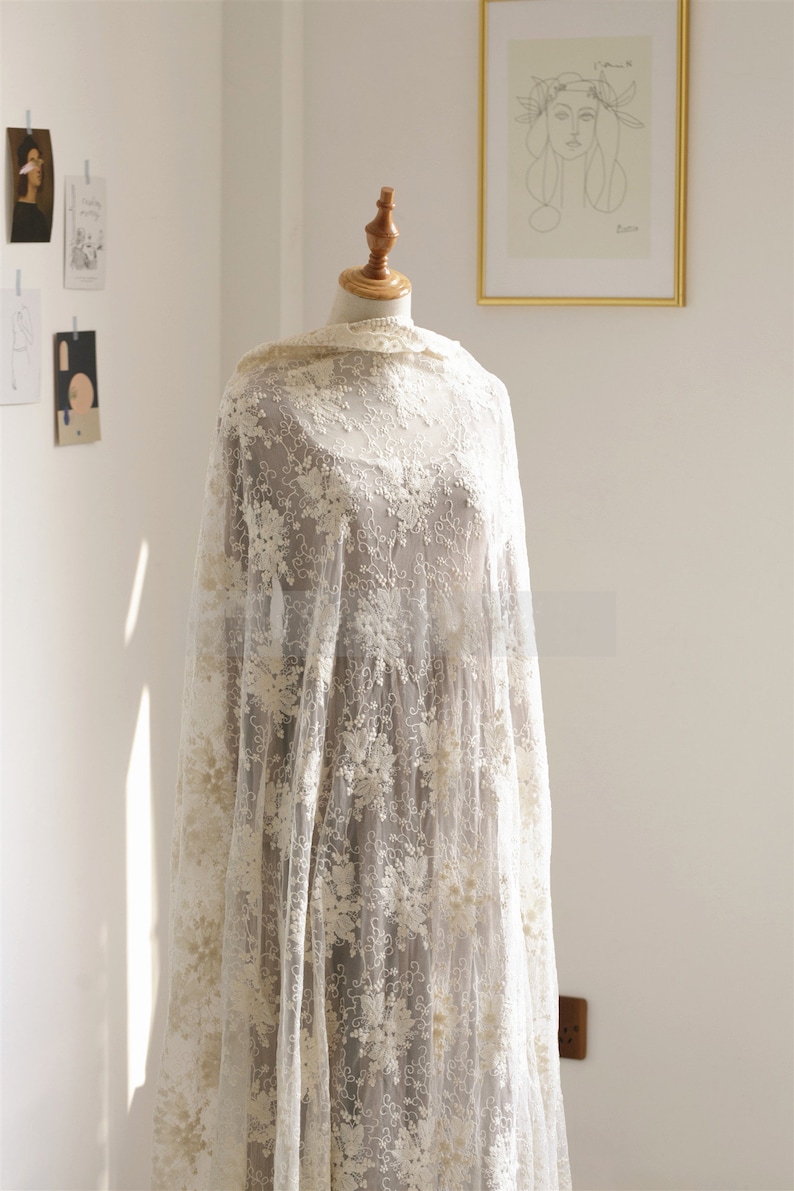May include: A white lace fabric with a delicate floral pattern. The fabric is draped over a mannequin and has a soft, flowing texture.
