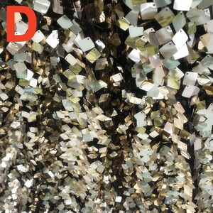 May include: Close-up of a fabric with a black background covered in small, square, iridescent sequins. The sequins reflect gold, silver, and pearlescent colors, creating a shimmering effect. The letter "D" is in the upper left corner.