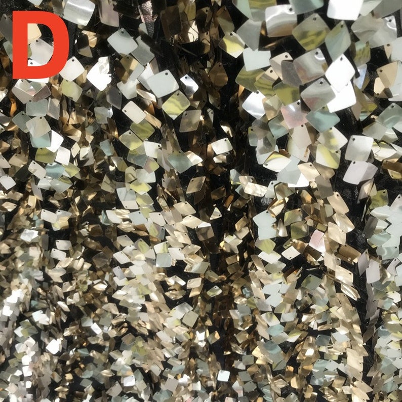 May include: Close-up of a fabric with a black background covered in small, square, iridescent sequins. The sequins reflect gold, silver, and pearlescent colors, creating a shimmering effect. The letter "D" is in the upper left corner.