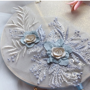 Puede incluir: Un delicado diseño de bordado floral con dos flores azul claro con centros blancos, rodeadas de hojas y tallos plateados. El bordado está sobre un fondo blanco.