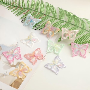 May include: A selection of embroidered butterfly appliques in pastel colours. The butterflies are displayed on a white surface with green fern leaves. Each butterfly features delicate threadwork and intricate details, ideal for crafting and embellishments.
