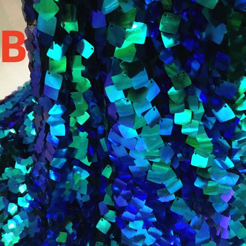 May include: Close-up of a fabric covered in square sequins. The sequins are in shades of blue and green, creating a shimmering effect. The letter "B" is visible in the upper left corner. The fabric appears to be draped.