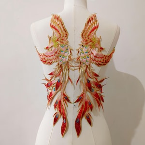 May include: Two embroidered phoenix patches. The patches feature a vibrant colour palette of red, gold, pink, and teal. The phoenixes have detailed wings and flowing tail feathers. The patches are likely for decorative purposes, such as embellishing clothing or accessories.