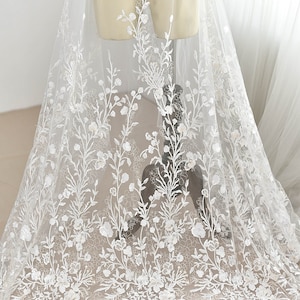 Pode incluir: Tecido de renda floral branca com um padrão delicado. A renda está sobre um fundo de tule transparente. O tecido é perfeito para vestidos de noiva, véus de noiva e outras roupas para ocasiões especiais.