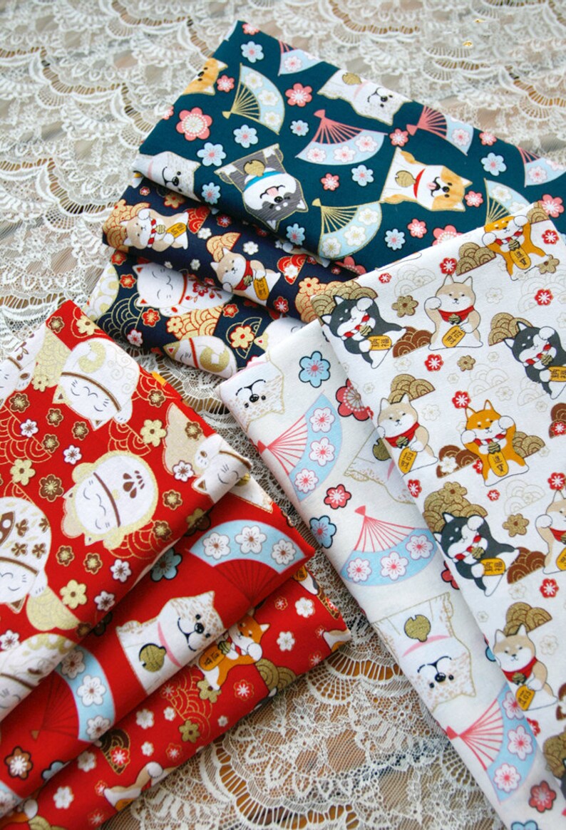 Puede incluir: Una pila de telas estampadas con dise&ntilde;os de inspiraci&oacute;n japonesa. Las telas muestran patrones repetidos de perros Shiba Inu, abanicos y motivos florales en rojo, azul marino y blanco. Las telas est&aacute;n dobladas y apiladas sobre una superficie de encaje.