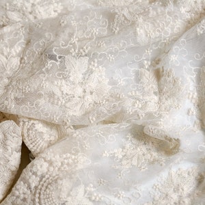 May include: White lace fabric with a delicate floral pattern. The fabric is soft and sheer, and it has a slightly textured surface.