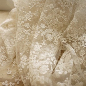 May include: Ivory white lace fabric with a delicate floral pattern. The lace is sheer and has a soft, romantic feel.