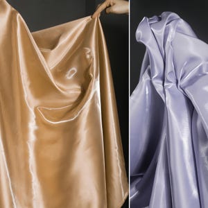 Op de afbeelding: Twee stukken stof, één goudkleurig en de andere lavendel, zijn gedrapeerd. De stof heeft een glanzende, satijnachtige uitstraling, met plooien en highlights die het licht vangen, waardoor een vloeiend effect ontstaat.