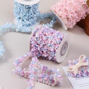 May include: Spools of decorative ribbon in pastel colors. The ribbon is a textured, looped design in shades of blue, pink, and purple. A small bow made from the ribbon sits on a white card.