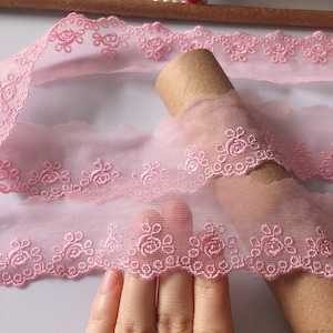 May include: Pink lace trim with a delicate floral pattern. The lace is soft and sheer, with a scalloped edge.