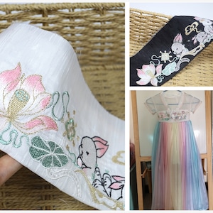 Könnte beinhalten: Weißer Stoffbesatz mit gestickten rosa und goldenen Lotusblumen, grünen Blättern und einem weißen Kaninchen. Der Besatz ist auf einem geflochtenen Korb zu sehen.