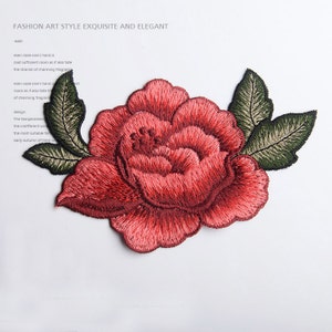 Könnte beinhalten: Eine rote gestickte Blume mit grünen Blättern. Die Blume ist eine einzelne Blüte mit einer gekräuselten Textur. Die Blätter sind mit einem hellgrünen Zentrum und dunkelgrünen Rändern detailliert.