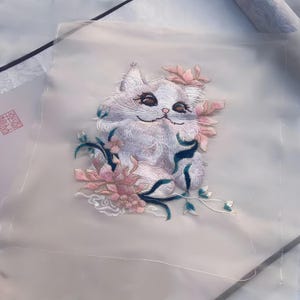 May include: Embroidered patch featuring a white cat with pink and blue floral accents. The cat has detailed facial features and is surrounded by delicate flowers and leaves. The embroidery is on a sheer, light-colored fabric, creating a soft, artistic effect.