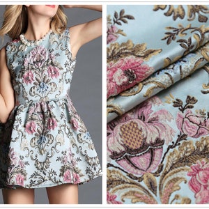 May include: A sleeveless, light blue dress with a floral pattern in pink, brown, and blue. The dress has a fitted bodice and a flared skirt. The fabric appears to be a brocade. A close-up shows the detailed floral design.