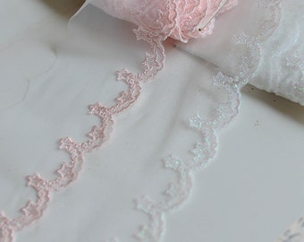 May include: Two rolls of delicate lace trim, one in pink and one in iridescent white. Both trims feature a scalloped edge with a repeating star and crescent moon design.