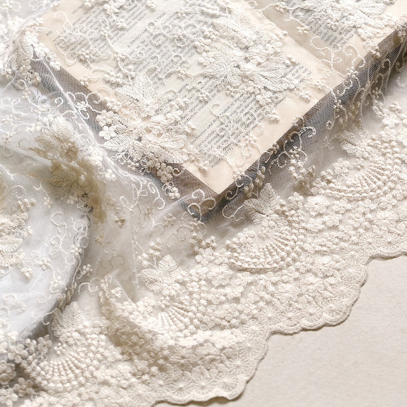 May include: White lace fabric with a delicate floral pattern. The lace is soft and sheer, with a scalloped edge.
