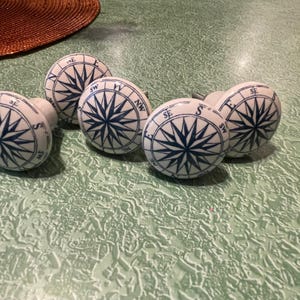 May include: A collection of five white ceramic cabinet knobs. Each knob features a blue compass rose design with directional indicators. The knobs are arranged on a textured, light green surface, suggesting a nautical or coastal theme.