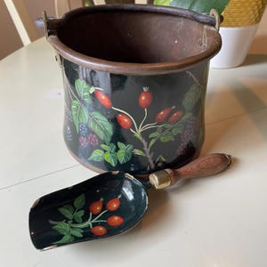 May include: A black metal bucket and matching scoop with a floral design. The bucket has a dark brown rim and handles. The scoop has a wooden handle and brass fitting. The floral design features red berries and green leaves.