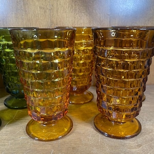 May include: Amber-colored vintage drinking glasses with a textured, geometric pattern. The glasses have a pedestal base and a flared rim. The glasses are arranged on a wooden surface. The glasses are a warm, inviting color.