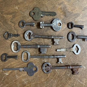 May include: A collection of antique keys in various shapes and sizes, arranged on a wooden surface. The keys are made of metal and show signs of age and wear, with different head designs and keyhole shapes. Some keys are small, while others are larger.