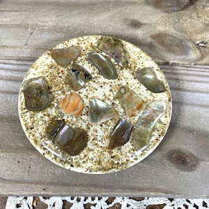 May include: A round coaster with a cream-colored base, speckled with small, light-brown particles. The coaster is adorned with various iridescent seashells and a single orange shell. The coaster is on a wooden surface.