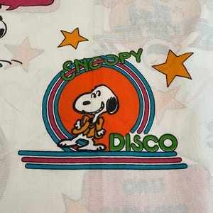 May include: White fabric with colorful Snoopy disco-themed graphics. The design includes Snoopy in a disco outfit, stars, and text that reads "Snoopy Disco" and "Boogie Down". Other text reads "The Dancing is Back!"