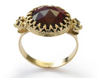 Könnte beinhalten: Ein goldener Ring mit einem großen, dunkelroten Edelstein, der in einer detaillierten, verzierten Goldfassung sitzt. Der Ring hat ein filigranes Design und ist mit kleineren, klaren Edelsteinen verziert.